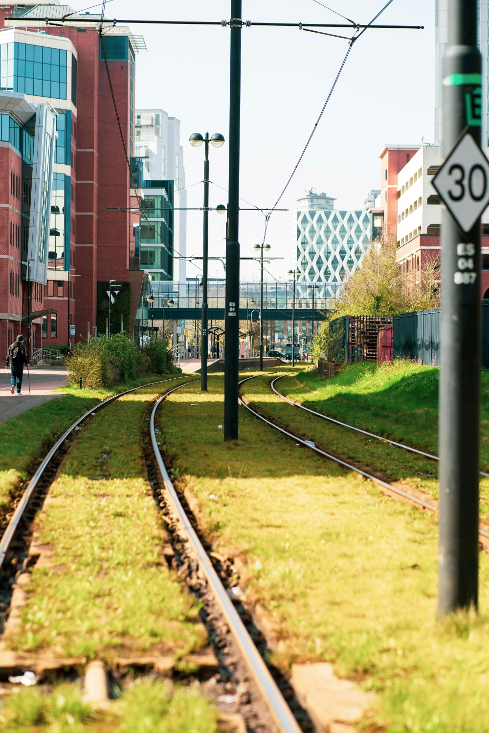 A tram track in GM covered with grass between the lines and Manchester's skyline in the background